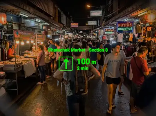 A crowded night market street seen from walking height, with a green arrow 100 metres ahead.